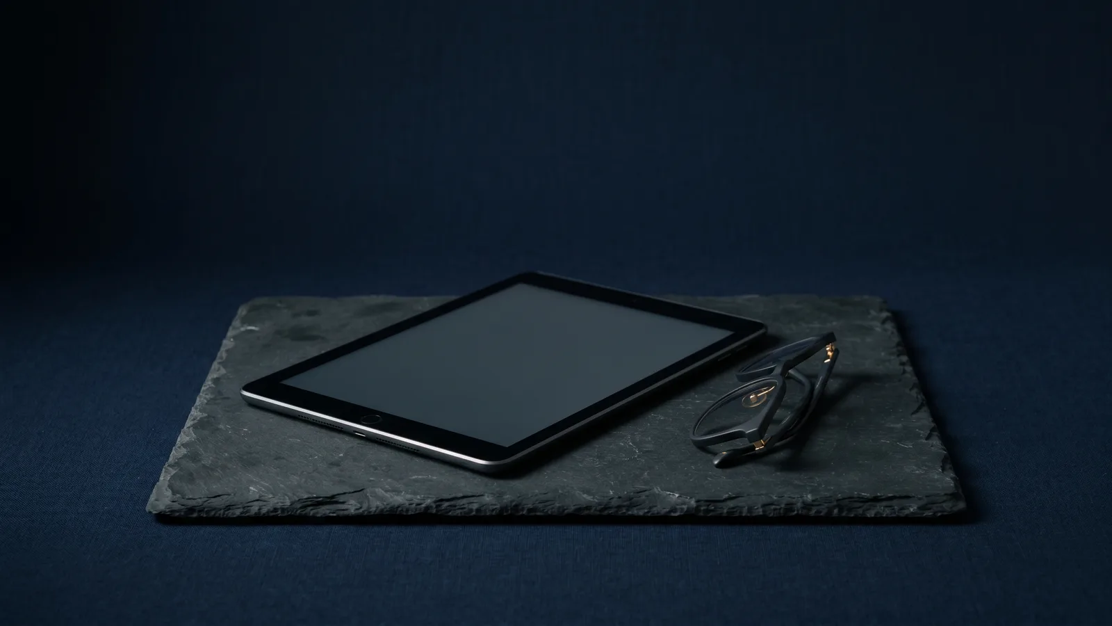 A tablet beside folded reading glasses on dark slate